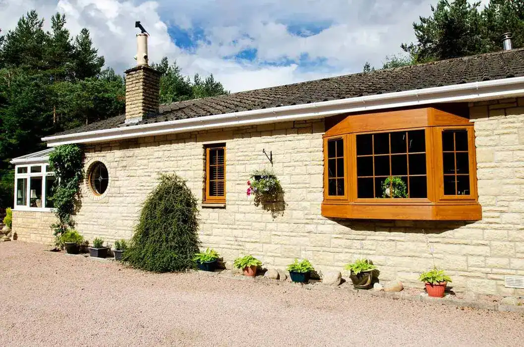 Stone bungalow with timber-framed bay window and double glazing, set against trees and a bright sky in Scotland.